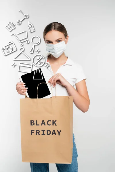 Mujer joven en máscara médica poner tableta digital con pantalla en blanco en bolsa de compras con letras de viernes negro cerca de la ilustración en blanco - foto de stock