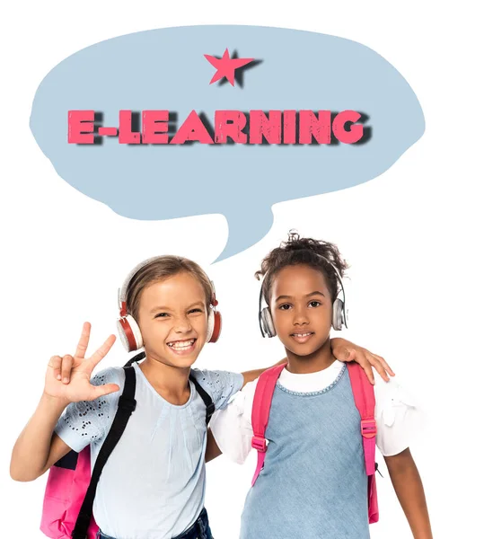 Schoolkid listening music in wireless headphones, hugging african american friend and showing three fingers near e-learning lettering on white — Stock Photo