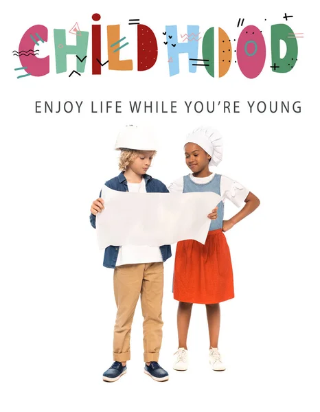 Curly boy in safety helmet and american kid in chef hat looking at blueprint near childhood, enjoy life while you are young lettering on white — Stock Photo