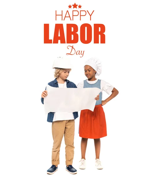 Curly boy in safety helmet and american kid in chef hat looking at blueprint near happy labor day lettering on white — Stock Photo