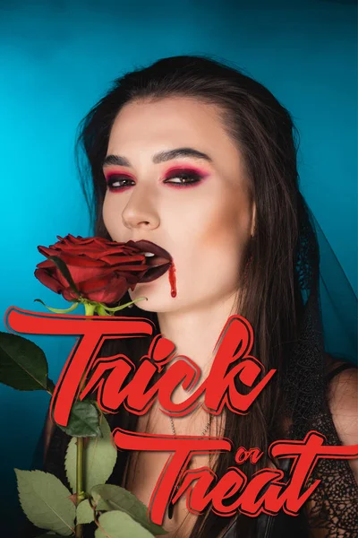 Mujer joven y espeluznante con sangre en la cara cerca de la rosa roja y engañar o tratar las letras en azul - foto de stock
