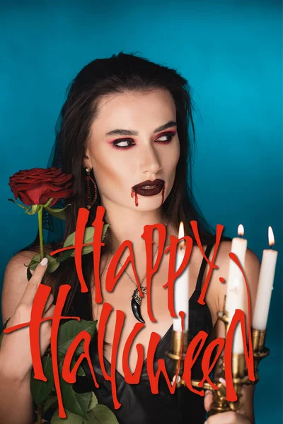 Junge Frau mit Blut im Gesicht bei brennenden Kerzen, Rose und fröhlichem Halloween-Schriftzug auf blau — Stockfoto