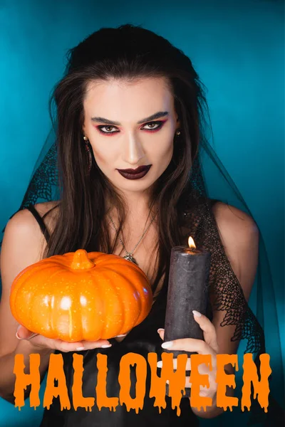 Femme pâle avec maquillage noir, voile tenant bougie brûlante et citrouille près de lettrage halloween sur bleu — Photo de stock