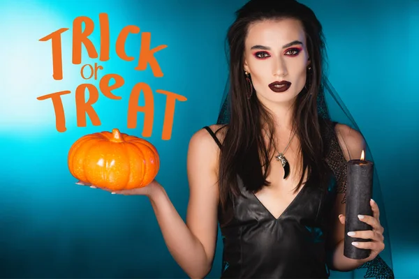 Young pale woman with black makeup and veil holding candle and pumpkin near trick or treat on blue — Stock Photo