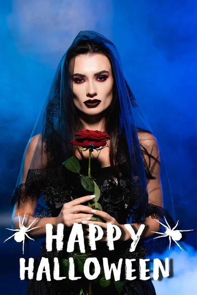 Brunette bride in black dress and veil holding red rose near happy halloween lettering on blue — Stock Photo
