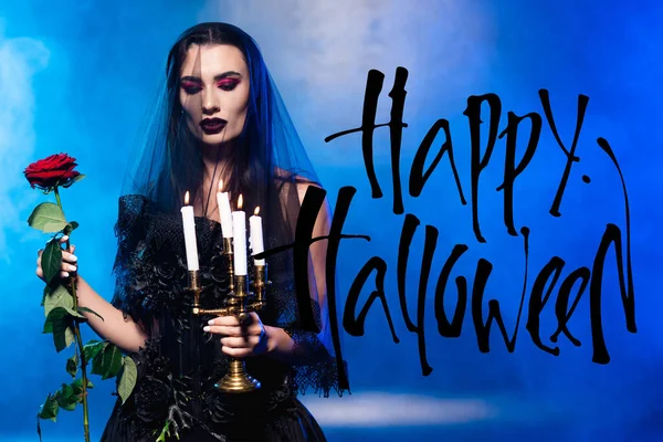 Woman in black dress and veil holding red rose and candles near happy halloween lettering on blue with smoke — Stock Photo