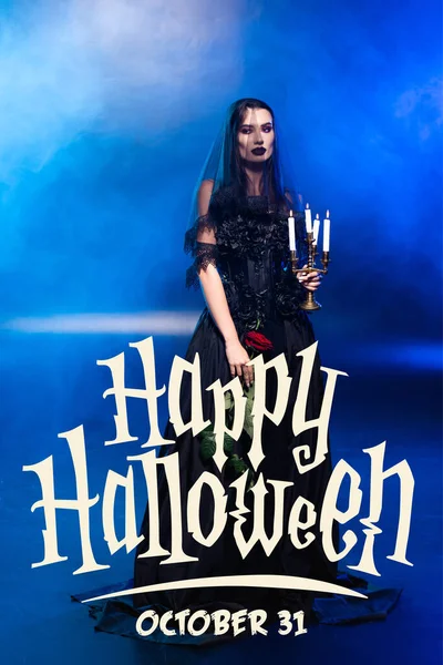 Bride in black dress and veil holding rose and candles near happy halloween lettering on blue with smoke — Stock Photo