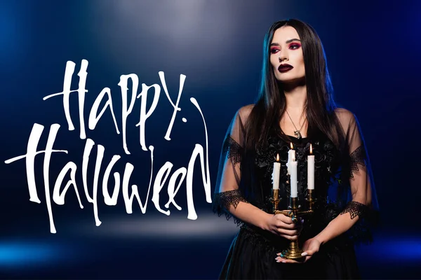 Femme en robe noire et voile tenant des bougies brûlantes près de joyeux lettrage halloween sur bleu — Photo de stock