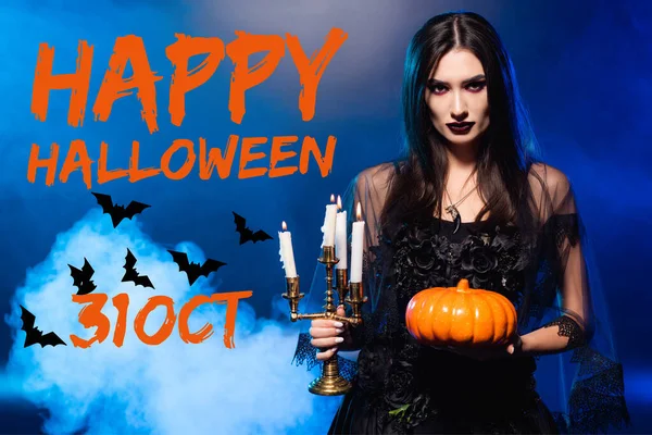 Mujer pálida con maquillaje aterrador sosteniendo calabaza y velas ardientes cerca de letras halloween feliz en azul con humo - foto de stock