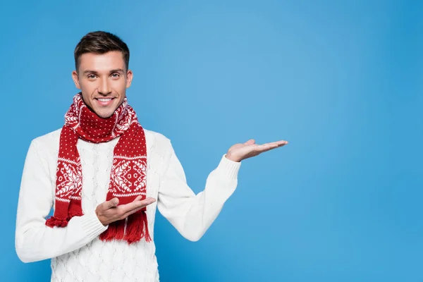 Smiling young adult man in sweater and scarf pointing with hands isolated on blue — Stock Photo