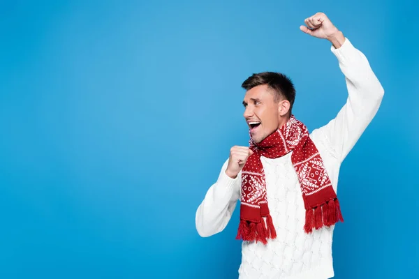 Excited young adult man with hands in air, wearing knitted sweater and scarf, looking away isolated on blue — Stock Photo