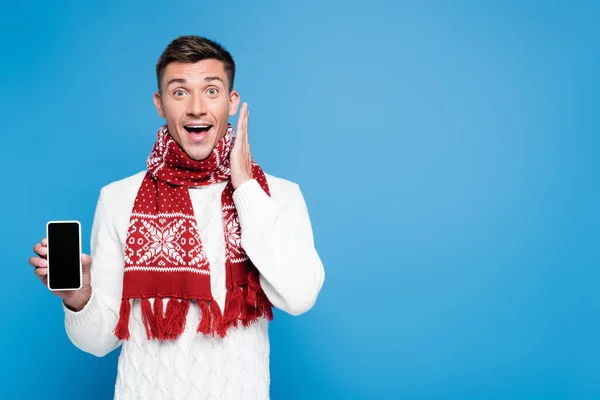 Excited young adult man with hand near head, showing smartphone with blank screen isolated on blue — Stock Photo