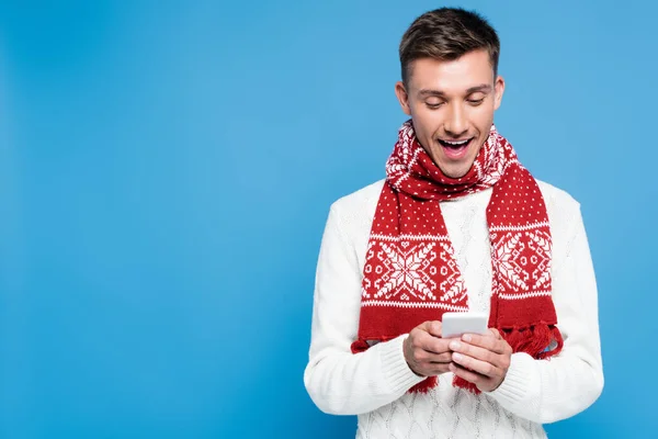 Emocionado hombre con bufanda de punto y suéter, mensajes de texto en el teléfono inteligente aislado en azul - foto de stock