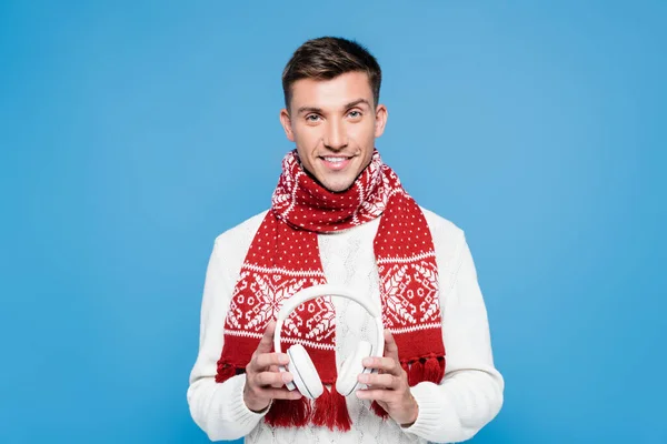 Uomo sorridente, indossa maglione e sciarpa, tenendo cuffie wireless bianche isolate su blu — Foto stock