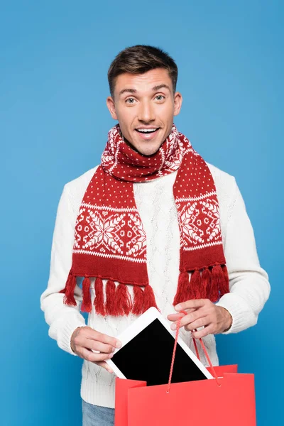 Smiling young adult man, putting digital tablet in red paper bag, while looking at camera isolated on blue — Stock Photo