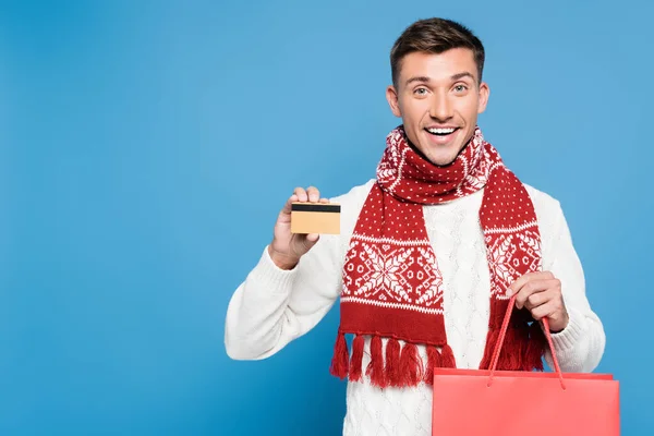 Sonriente hombre adulto joven mostrando tarjeta de crédito, sosteniendo bolsa de papel rojo, mientras mira la cámara aislada en azul - foto de stock