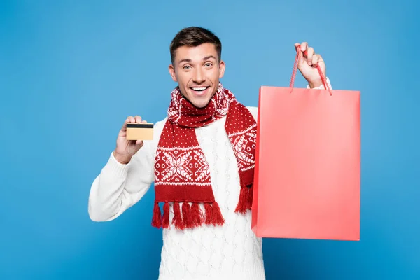 Feliz joven adulto mostrando tarjeta de crédito y bolsa de papel en blanco aislado en azul - foto de stock