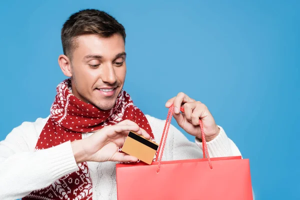 Portrait of smiling young adult man, putting credit card in red paper bag isolated on blue — Stock Photo