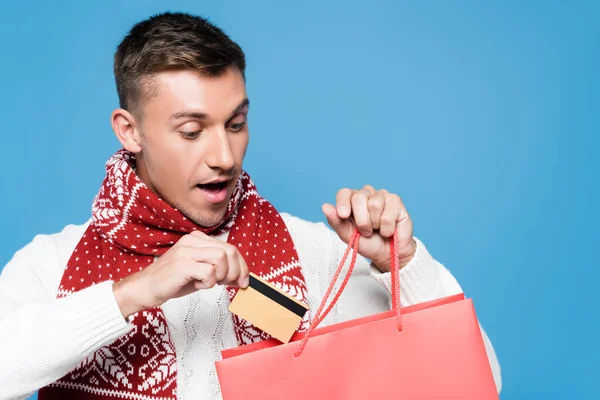 Retrato del joven adulto sorprendido, colocando tarjeta de crédito en bolsa de papel roja aislada en azul - foto de stock