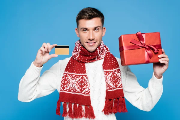 Uomo sorridente che indossa sciarpa e maglione, mostrando regalo avvolto e carta di credito isolata su blu — Foto stock