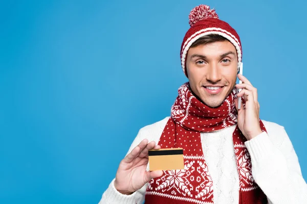 Hombre feliz en gorra de punto hablando en el teléfono inteligente y mostrando la tarjeta de crédito aislado en azul - foto de stock