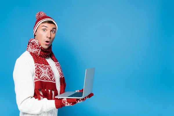 Überraschter Mann in warmer Kleidung, Laptop in der Hand, während er isoliert auf blauem Grund in die Kamera blickt — Stockfoto