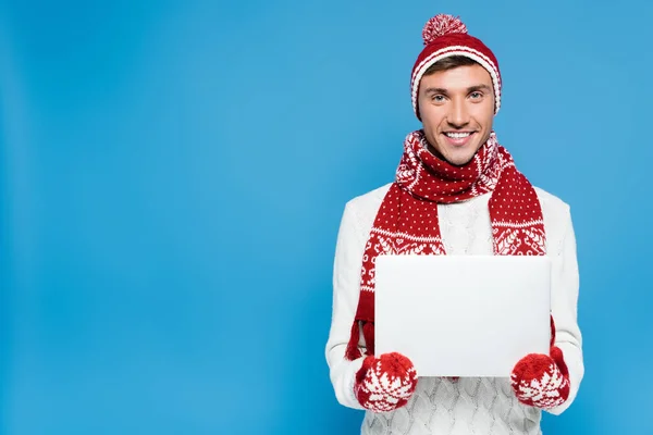 Вид спереду усміхненого молодого дорослого чоловіка в теплому одязі, що стоїть з ноутбуком ізольовано на синьому — стокове фото