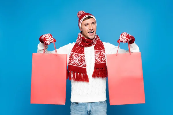 Happy young adult man in mittens holding red paper bags isolated on blue — Stock Photo