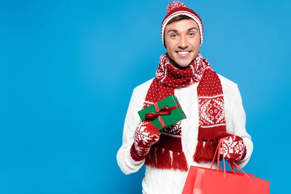 Souriant jeune homme adulte en vêtements chauds tenant petite boîte cadeau et sac en papier rouge isolé sur bleu — Photo de stock