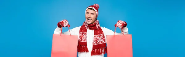 Cheerful young adult man in mittens holding red paper bags isolated on blue, banner — Stock Photo