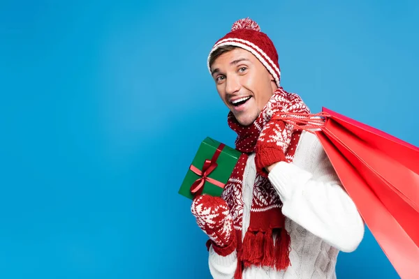Excited young adult man with red paper bags behind back, holding small wrapped present isolated on blue — Stock Photo
