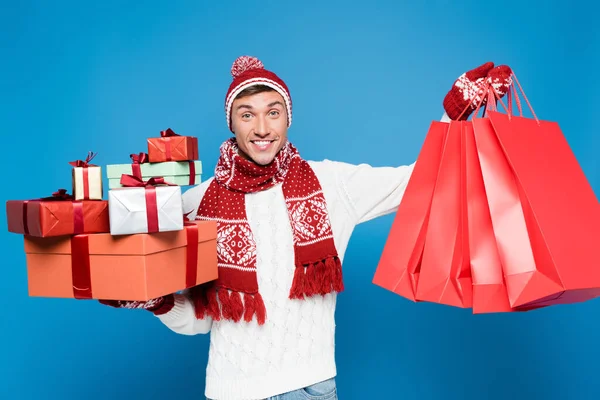 Front view of happy man in warm clothing holding bunch of gift boxes and red paper bags isolated on blue — Stock Photo