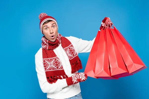 Hombre sorprendido en manoplas levantando bolsas de papel rojo, mientras mira a la cámara aislada en azul - foto de stock