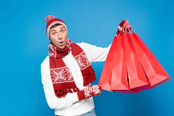 Hombre sorprendido en manoplas levantando bolsas de papel rojo, mientras mira a la cámara aislada en azul - foto de stock