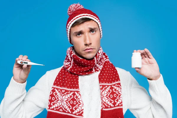 Portrait of sick man in knitted scarf and hat showing thermometer and jar of tablets isolated on blue — Stock Photo