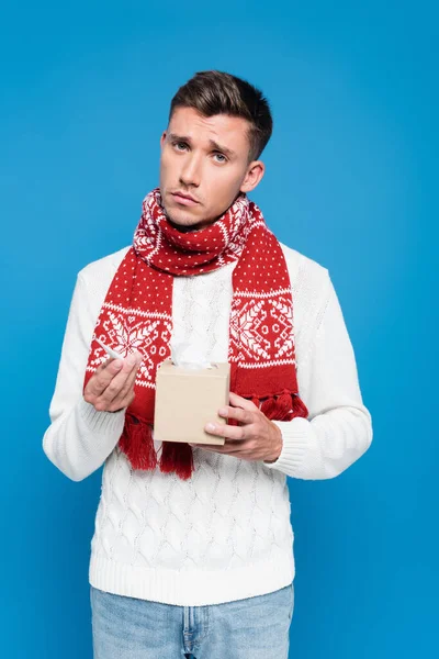 Kranker und verärgerter junger erwachsener Mann mit Thermometer und Schachtel Taschentücher isoliert auf blauem Grund — Stockfoto