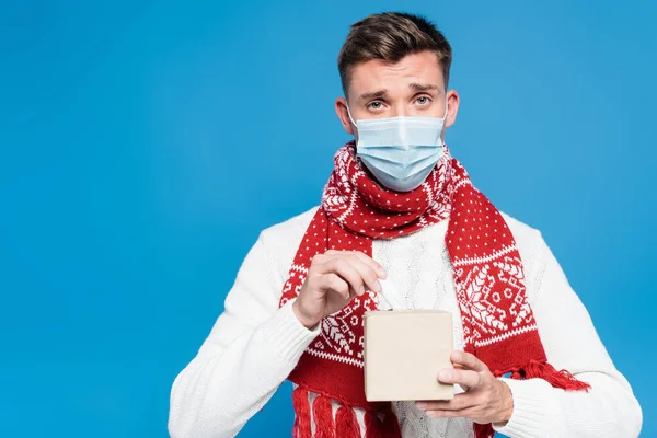 Молодой взрослый мужчина в медицинской маске держит коробку с тканями, изолированными на голубом — стоковое фото
