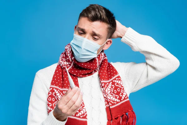 Hombre adulto joven con la mano cerca de la cabeza, con máscara médica y mirando el termómetro aislado en azul - foto de stock