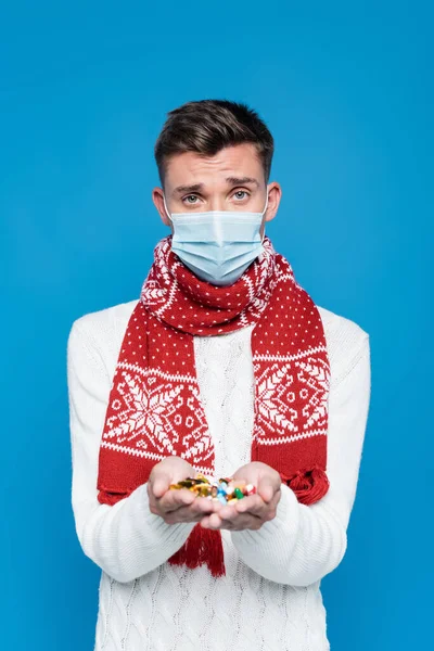 Homme portant un masque médical, debout avec une poignée de pilules isolées sur bleu — Photo de stock