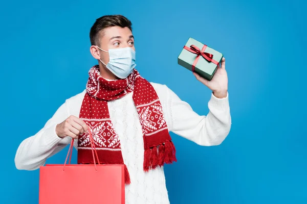 Hombre adulto joven con máscara médica, sosteniendo una pequeña caja de regalo y una bolsa de papel roja, mientras mira hacia otro lado aislado en azul - foto de stock