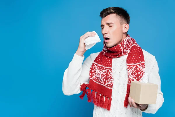 Молодий дорослий чоловік в червоному шарфі чхає, тримаючи коробку тканин ізольовано на синьому — Stock Photo