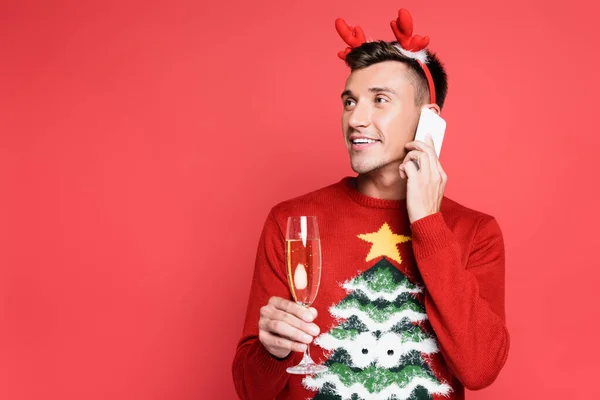 Homme souriant en pull avec pin parlant sur smartphone et tenant un verre de champagne isolé sur rouge — Photo de stock