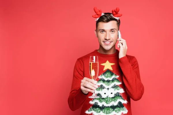 Homme souriant en pull de Noël avec verre de champagne parlant sur smartphone sur fond rouge — Photo de stock