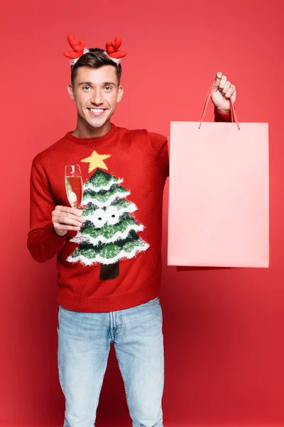 Homme souriant en pull avec pin tenant un verre de champagne et un sac à provisions sur fond rouge — Photo de stock