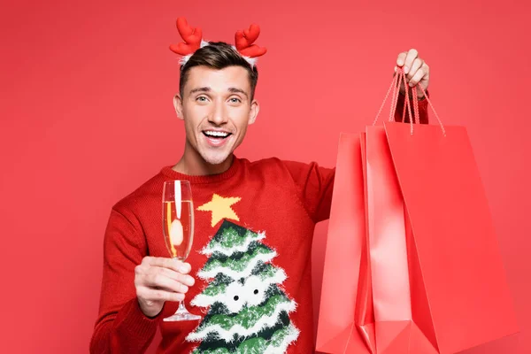 Uomo sorridente in maglione di Natale che tiene un bicchiere di champagne e borse della spesa isolate su rosso — Foto stock