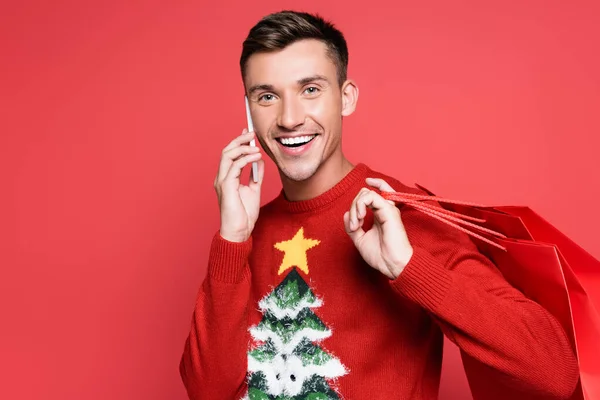 Hombre alegre en jersey navideño hablando en smartphone y sosteniendo bolsas aisladas en rojo - foto de stock