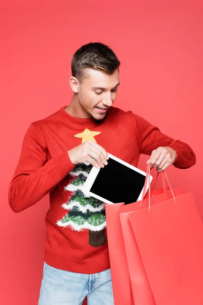 Homme positif en pull avec pin mettre tablette numérique dans des sacs à provisions isolés sur rouge — Photo de stock