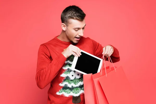 Hombre excitado en jersey navideño poniendo tableta digital en bolsas aisladas en rojo - foto de stock