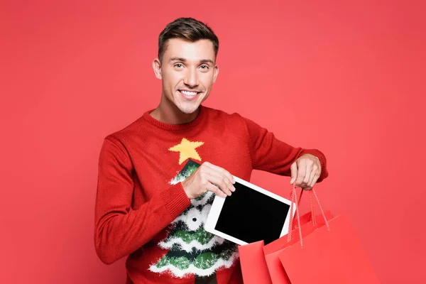 Uomo sorridente in maglione con albero di Natale mettere tablet digitale nella shopping bag isolato su rosso — Foto stock