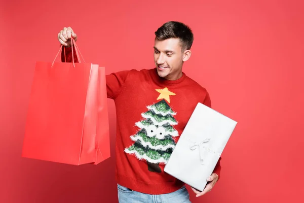Hombre sonriente en suéter con árbol de Navidad sosteniendo bolsas de compras y caja de regalo sobre fondo rojo - foto de stock
